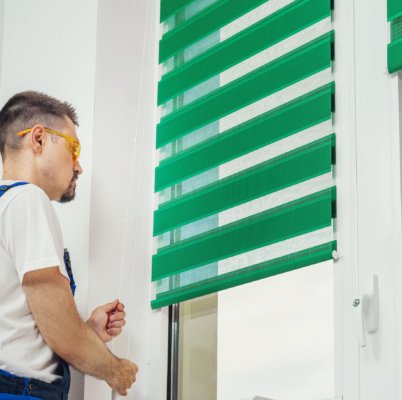 Young man standing in front of bright window in room adjusting green color blinds curtains, white painted walls, during removing, cleaning.